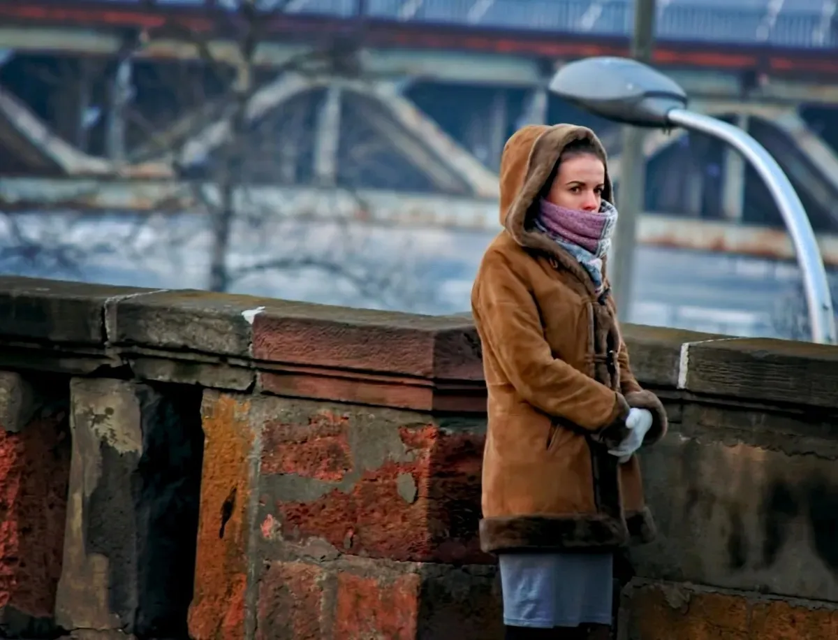 Woman in a brown coat leaning on a bridge railing